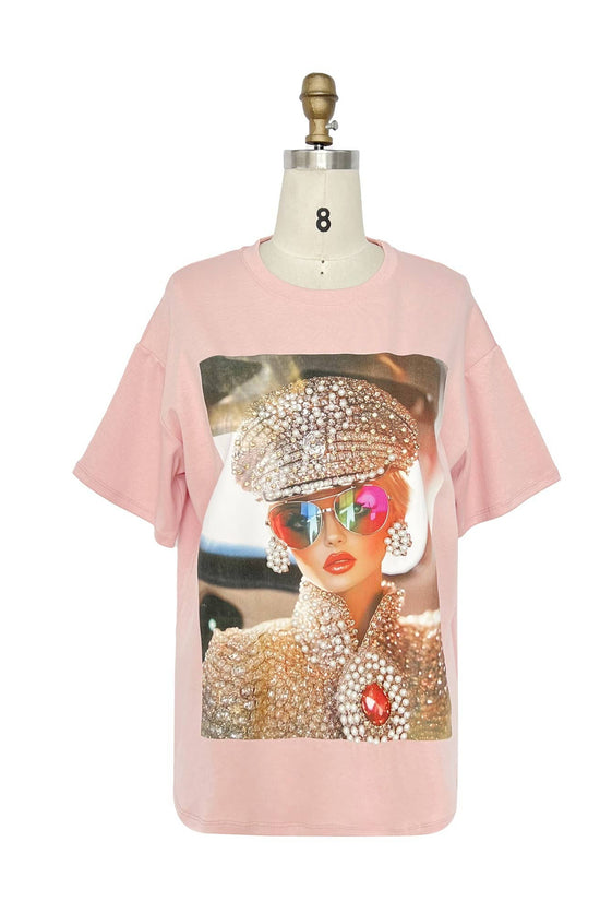 Pink t-shirt with a graphic of a person wearing a rhinestone hat and sunglasses on a mannequin against a white background. Born To Glam