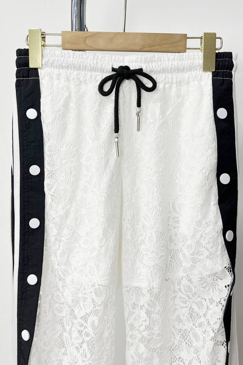 White lace pants with black polka dot straps on a hanger against a white background Born To Glam