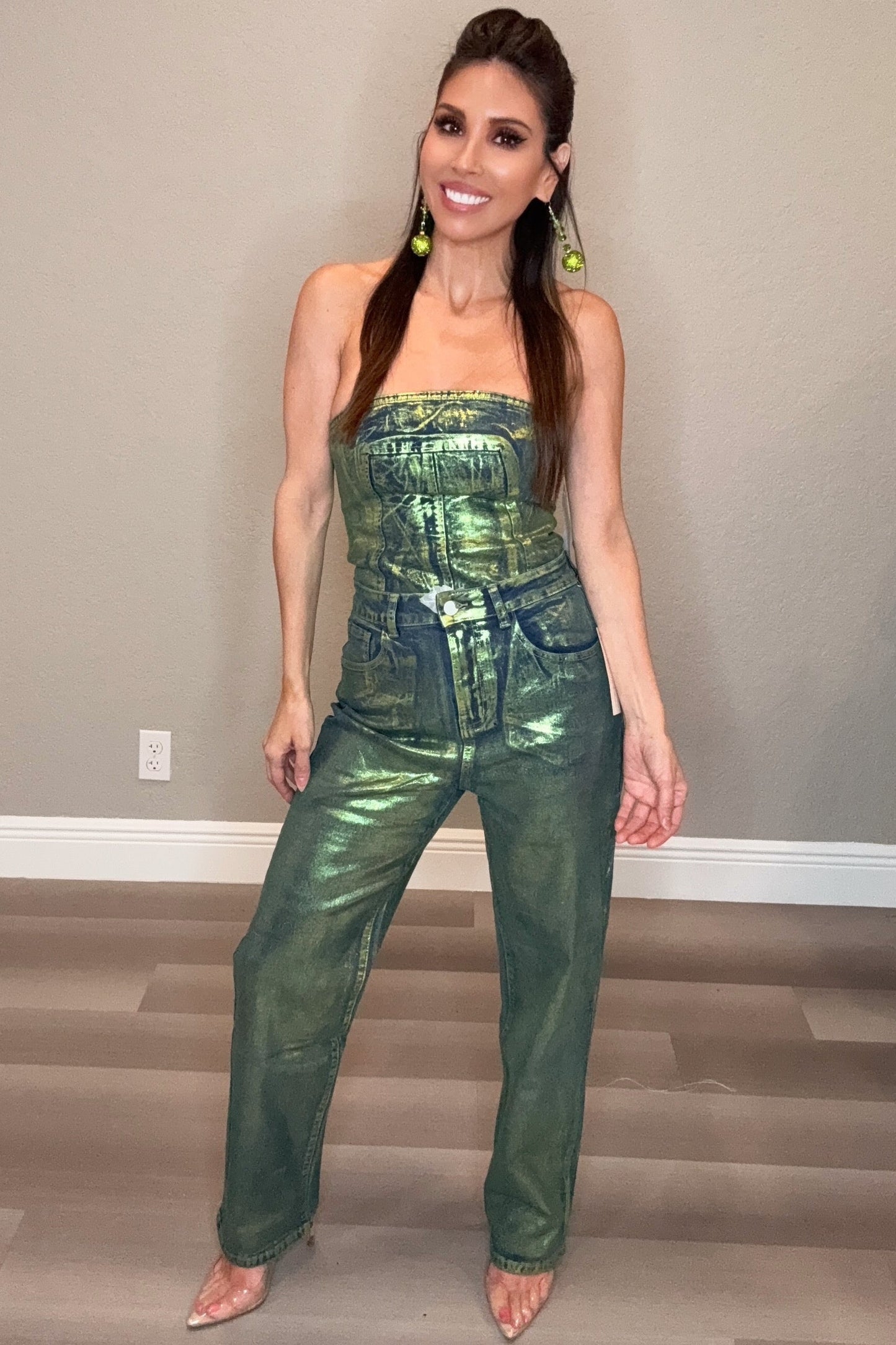 Woman wearing a green metallic jumpsuit standing in a room with a neutral background. Born To Glam