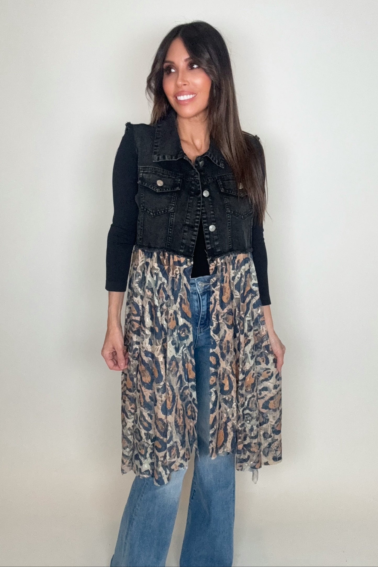 Woman wearing a black denim vest over a leopard print dress with blue jeans on a plain background Born To Glam