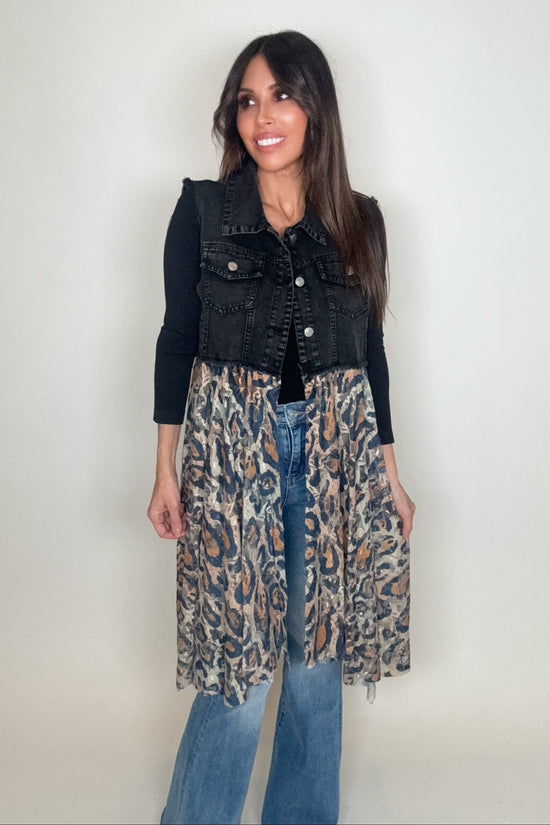 Woman wearing a black denim vest over a leopard print dress with blue jeans on a plain background Born To Glam