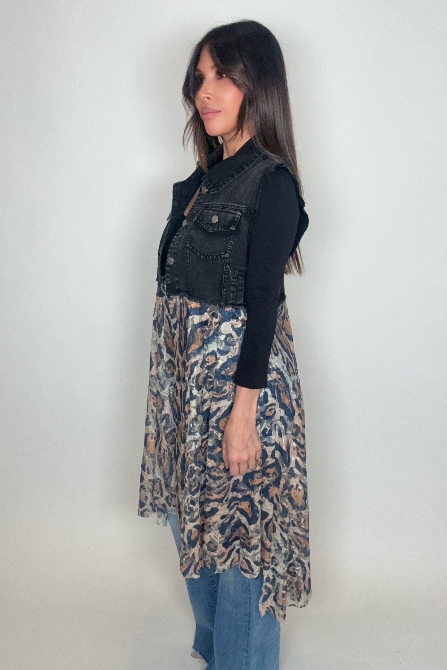 Woman wearing a black denim vest over a patterned dress with blue jeans on a white background Born To Glam