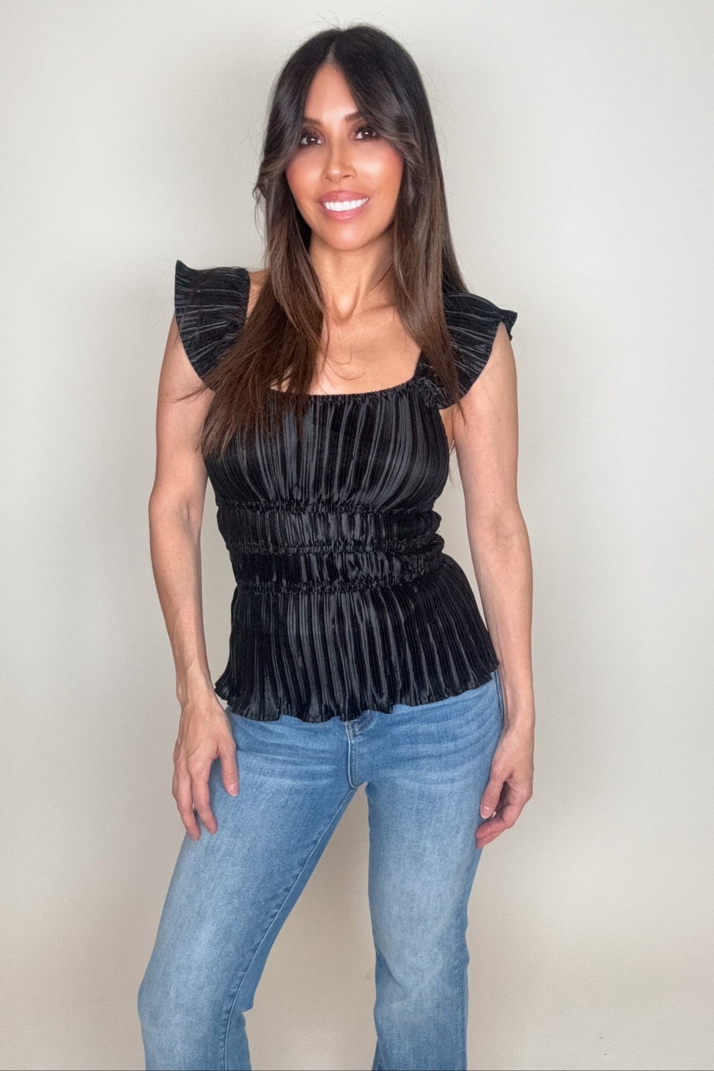 Woman wearing a black pleated top and blue jeans against a plain background Born To Glam