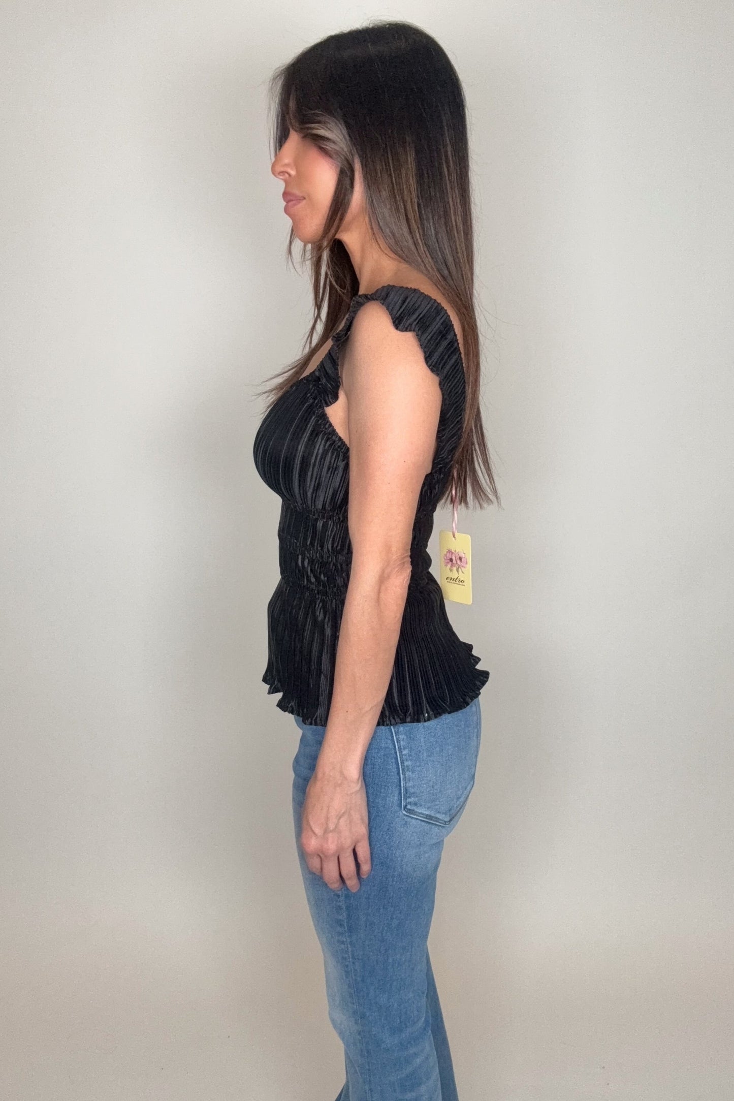 Woman wearing a black sleeveless top with a visible tag against a plain background Born To Glam