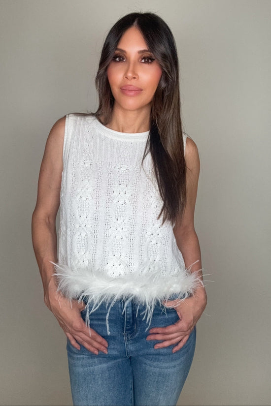 Woman wearing a white sleeveless top with feather details and blue jeans against a neutral background Born To Glam