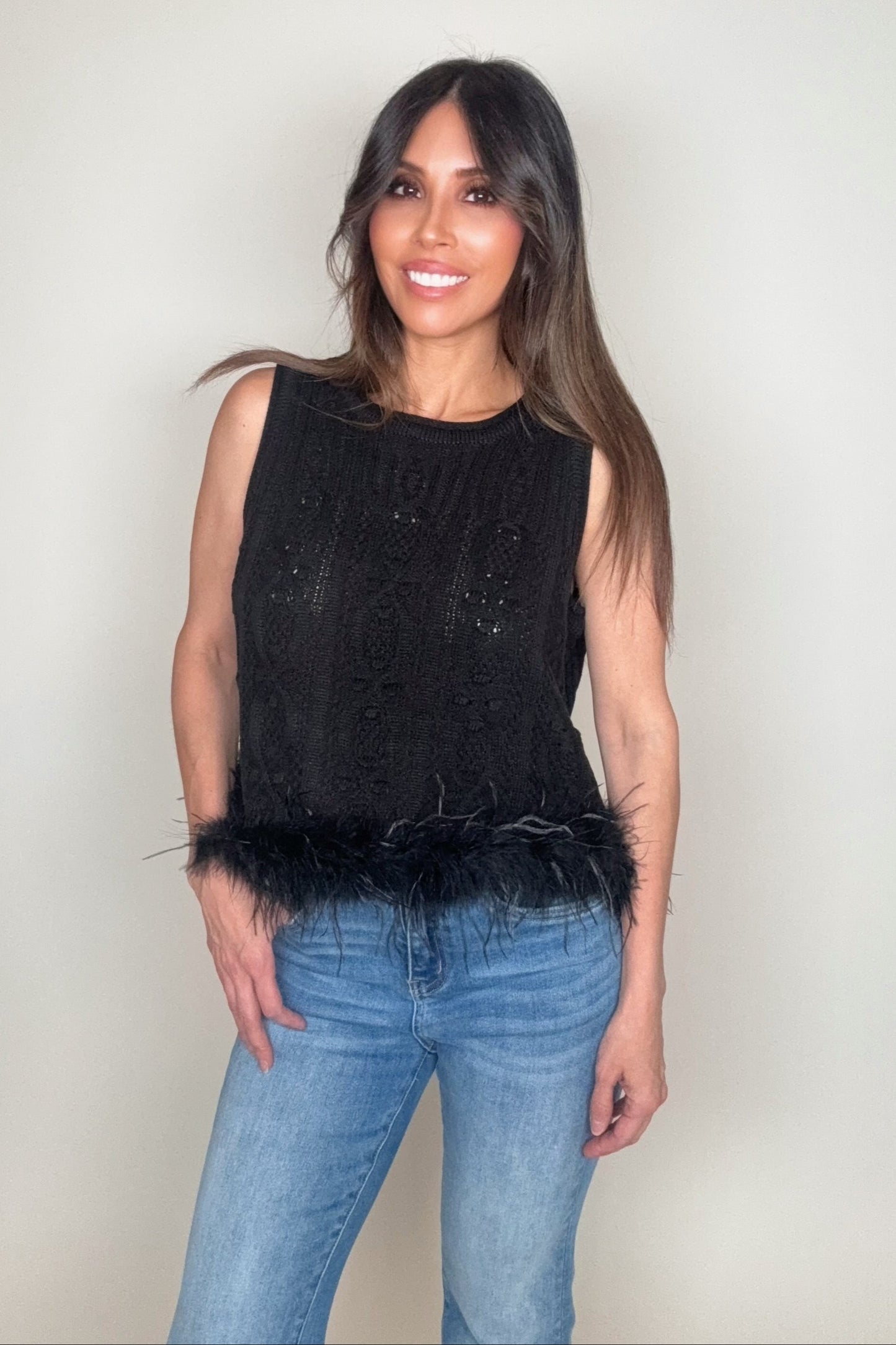 Woman wearing a black sleeveless top with feather details and blue jeans on a plain background Born To Glam
