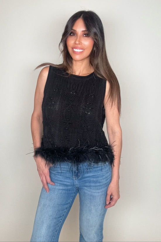 Woman wearing a black sleeveless top with feather details and blue jeans on a plain background Born To Glam