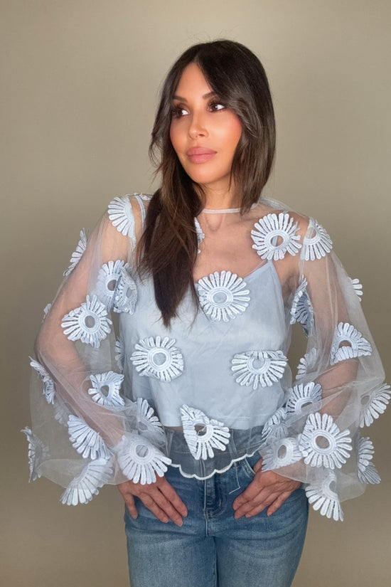 Woman wearing a light blue blouse with white floral patterns and sheer sleeves against a beige background Born To Glam