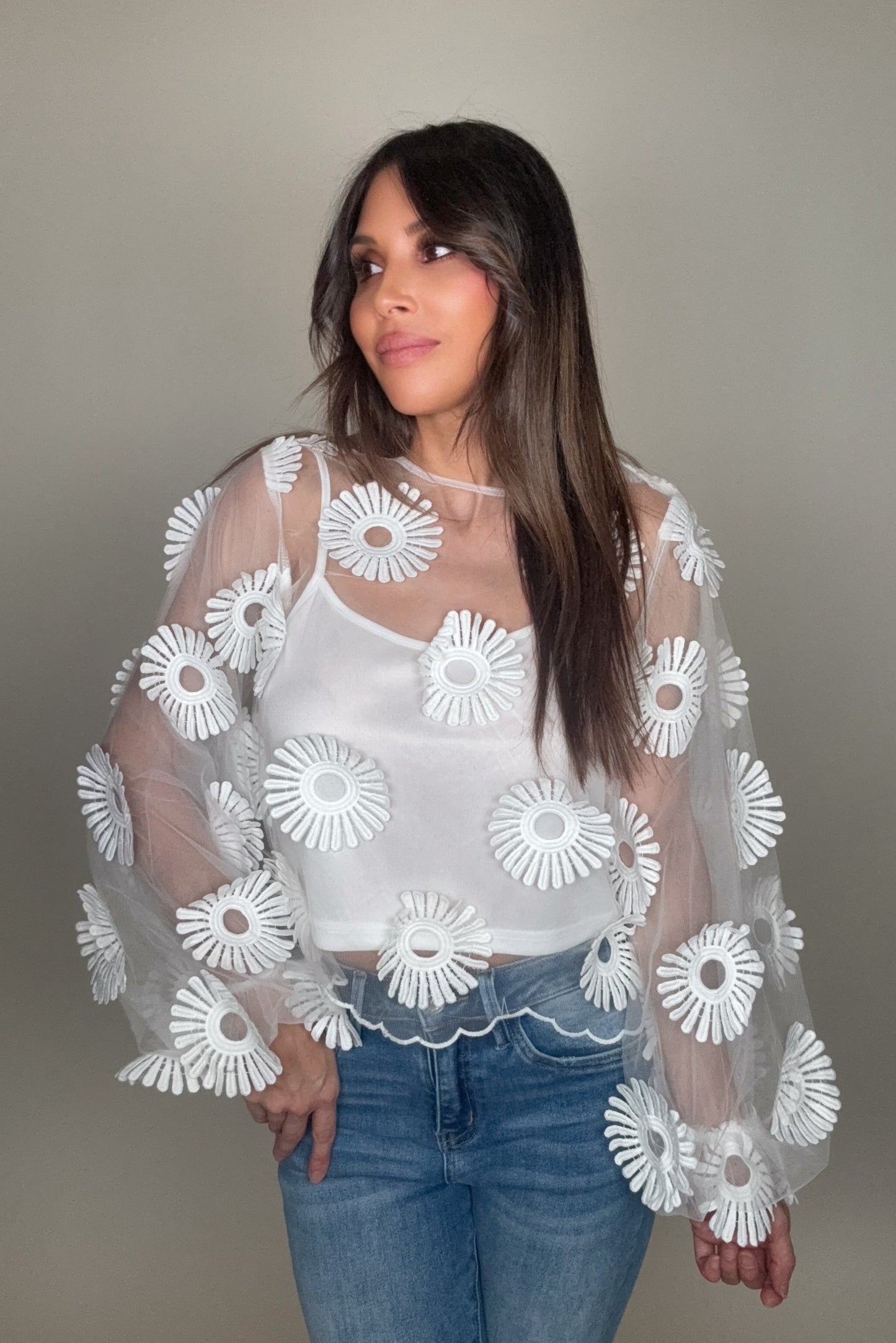 Woman wearing a white blouse with floral patterns and blue jeans against a neutral background Born To Glam