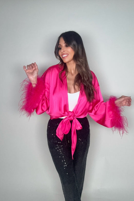 Woman wearing a bright pink satin robe with feather sleeves against a plain background Born To Glam