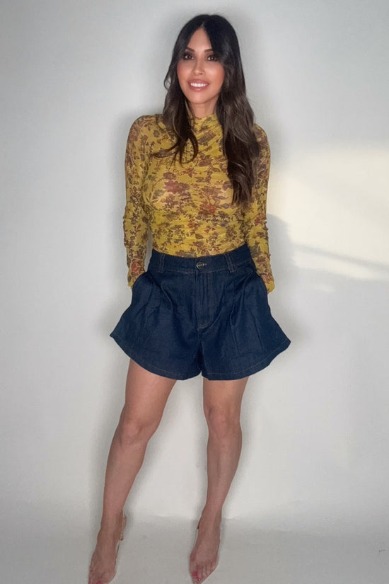 Woman wearing a yellow patterned top and dark blue shorts against a plain background Born To Glam