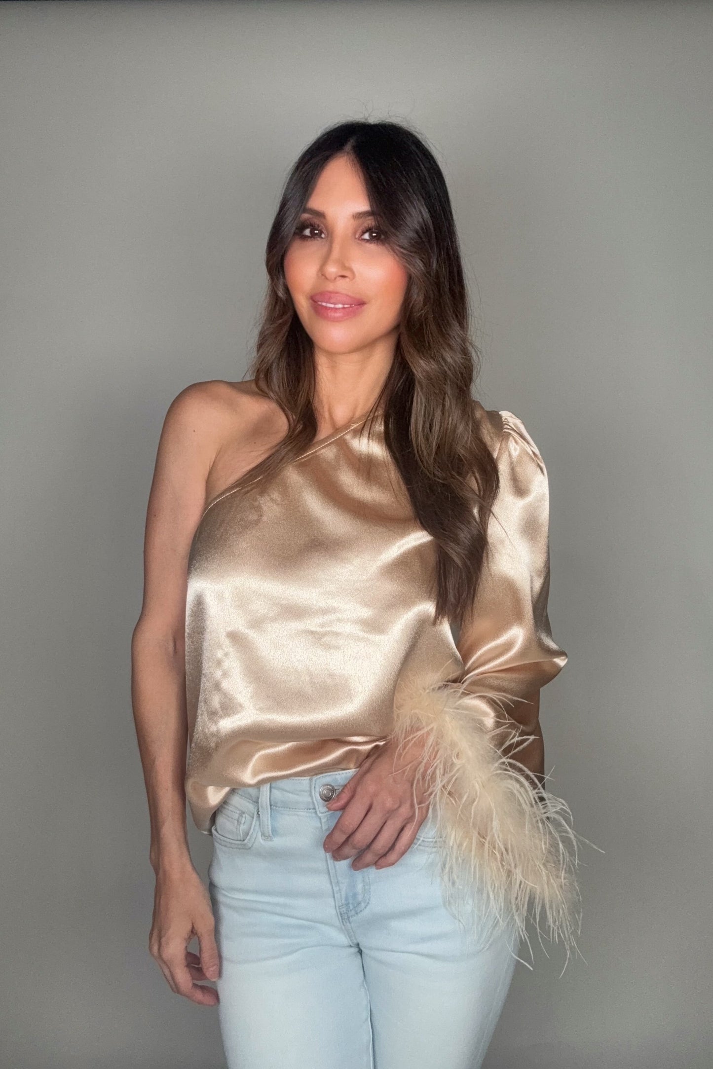 Woman wearing a gold satin top with feather details and light blue jeans against a gray background Born To Glam
