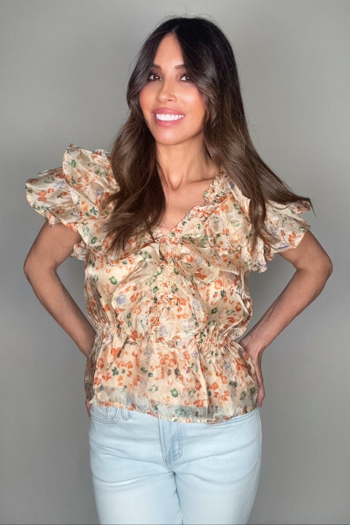 Woman wearing a floral blouse and light blue jeans against a gray background Born To Glam