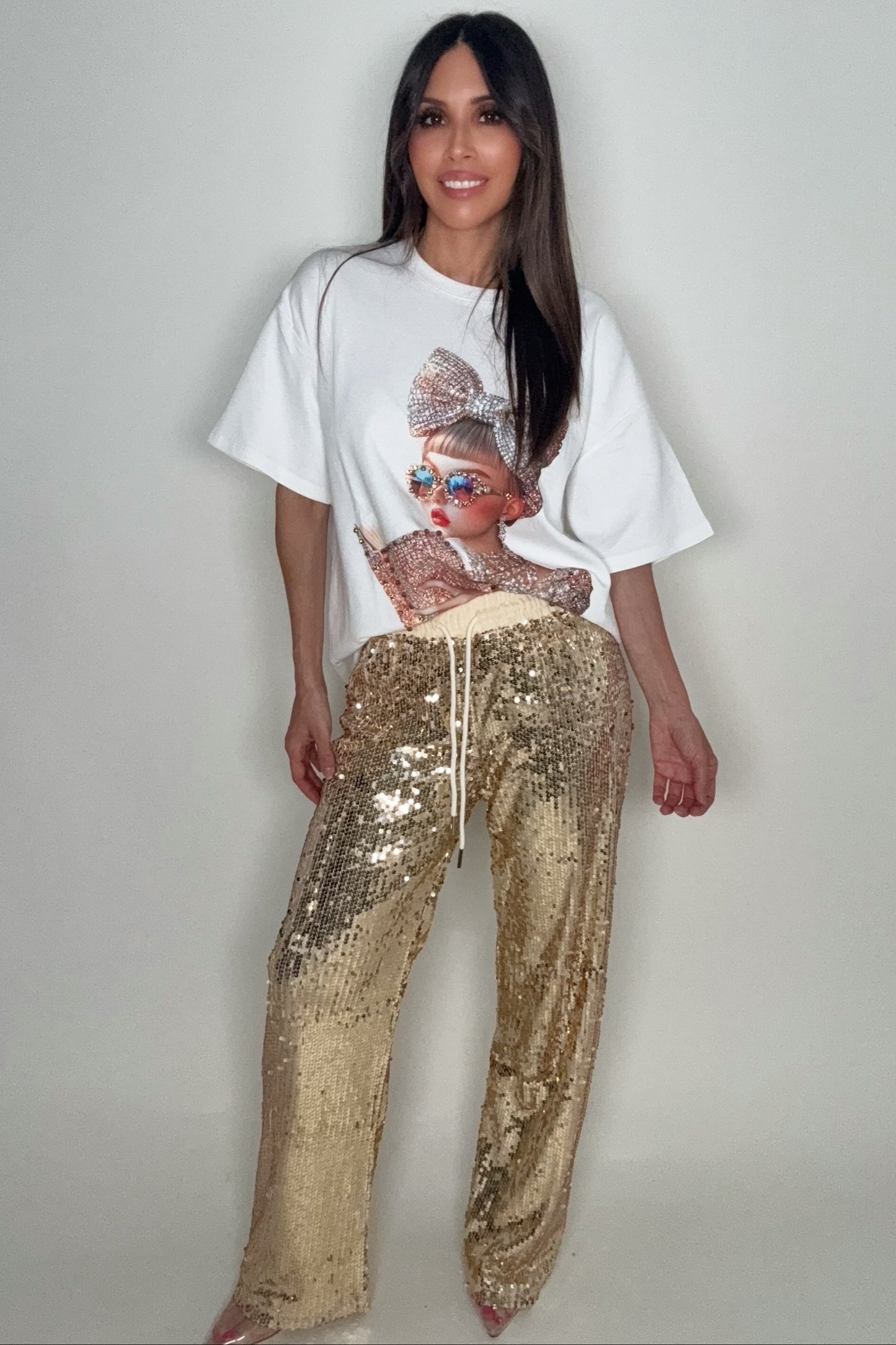 Woman wearing a white t-shirt with a graphic design and gold sequin pants on a plain background Born To Glam