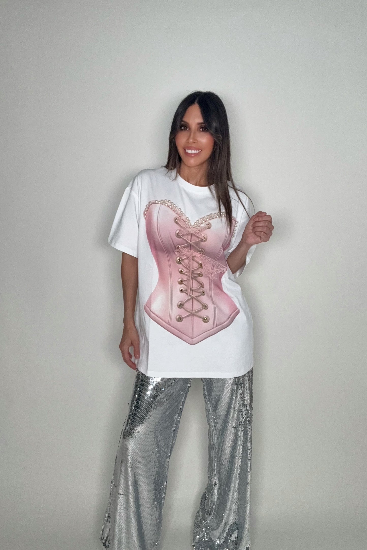Person wearing a white t-shirt with a pink corset design and silver sequin pants on a plain background Born To Glam