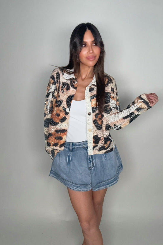 Woman wearing a leopard print jacket, white top, and denim shorts against a plain background Born To Glam
