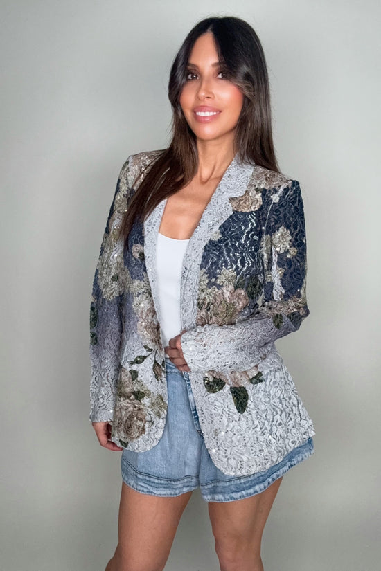 Woman wearing a floral blazer over a white top and denim shorts on a plain background Born To Glam