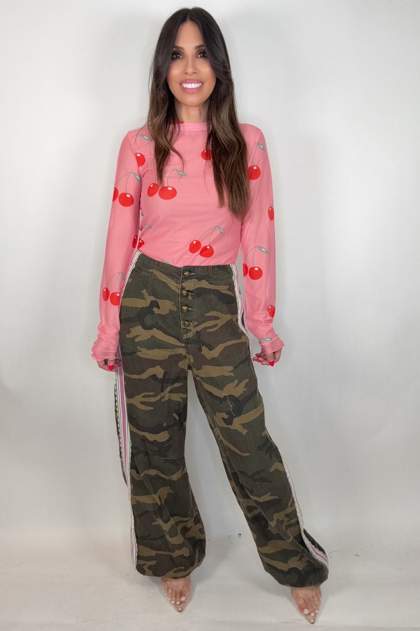 Woman wearing a pink long-sleeve top with red cherry pattern and camouflage pants with white stripes on a white background Born To Glam