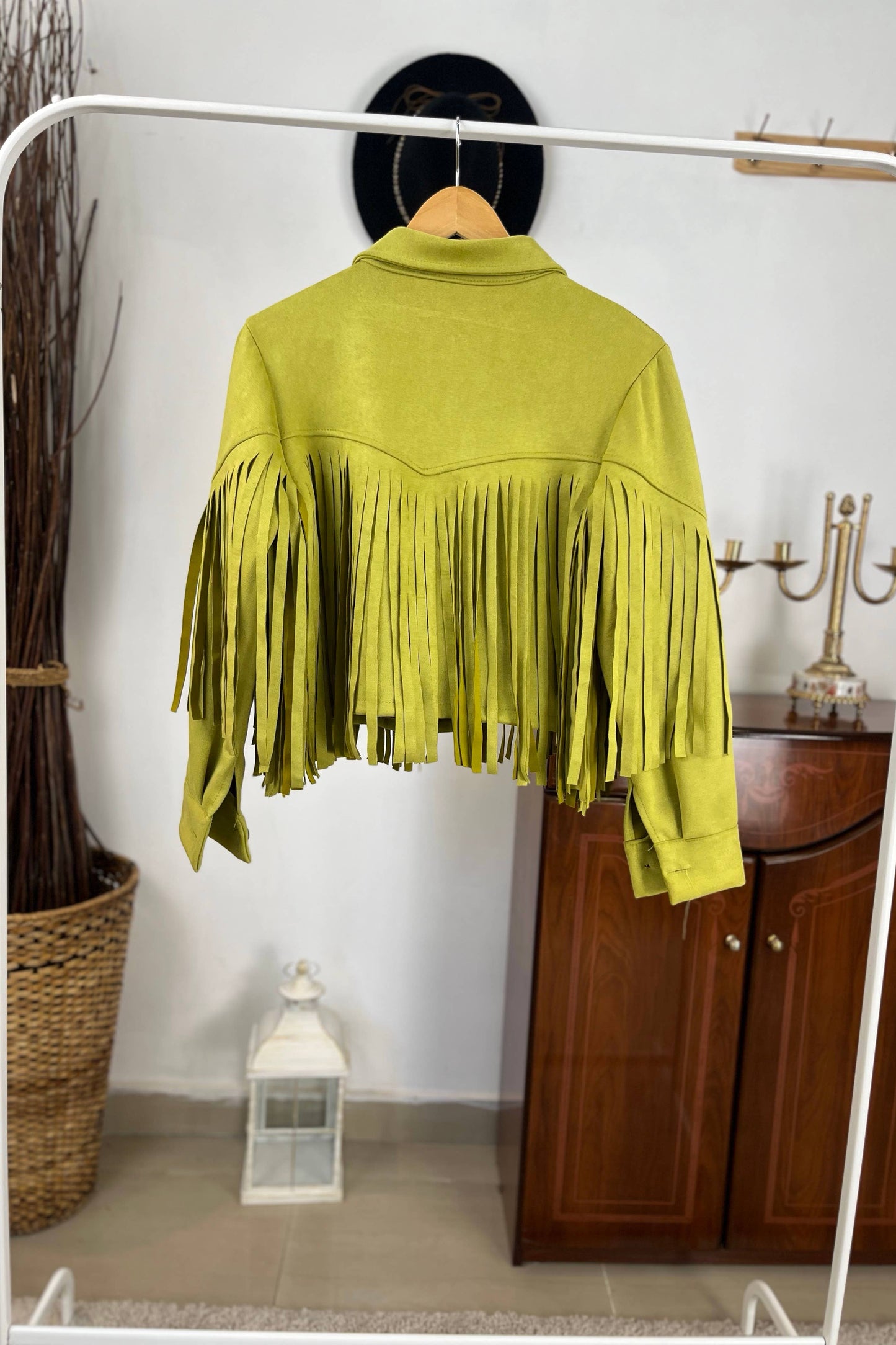 Green fringed jacket on a white rack with a wooden cabinet and decorative items in the background. Born To Glam
