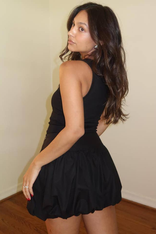 Woman wearing a black dress against a plain wall. Born To Glam