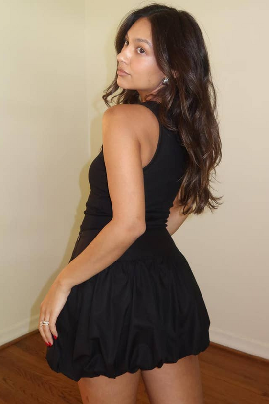 Woman wearing a black dress against a plain wall. Born To Glam